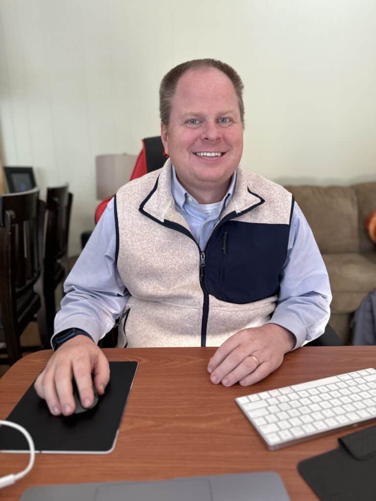 Image of a cheerful man sitting at a desk with a friendly smile, exuding a sense of contentment and professionalism. The man appears focused and confident, embodying the 'Best Effort" and giving his 100% in his demeanor.